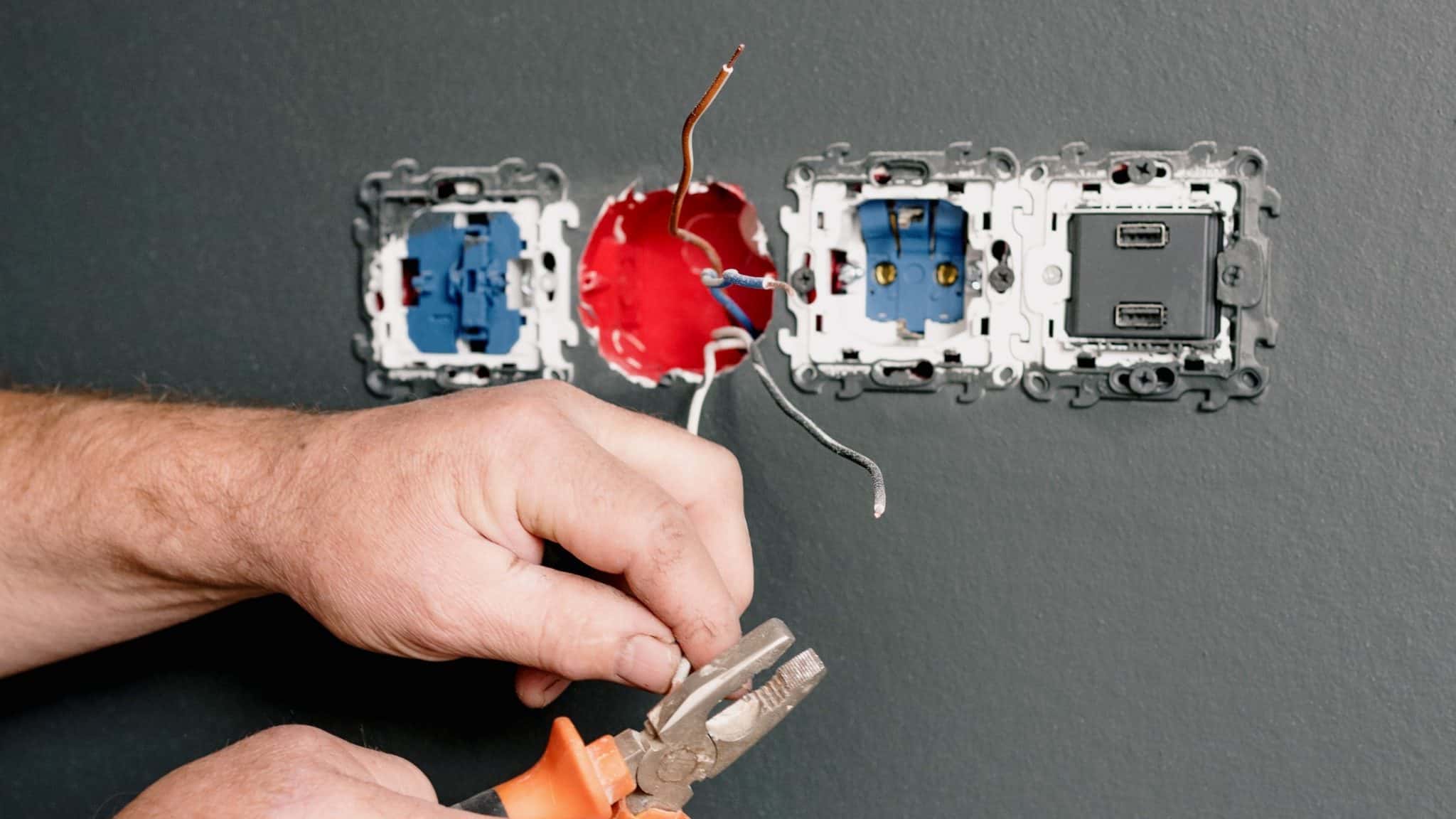 Electrician's hands holding pliers attaching wires to wall sockets on grey wall