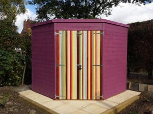 Pent roof shed in a corner on slabs painted purple with multi-coloured striped painted on locked door