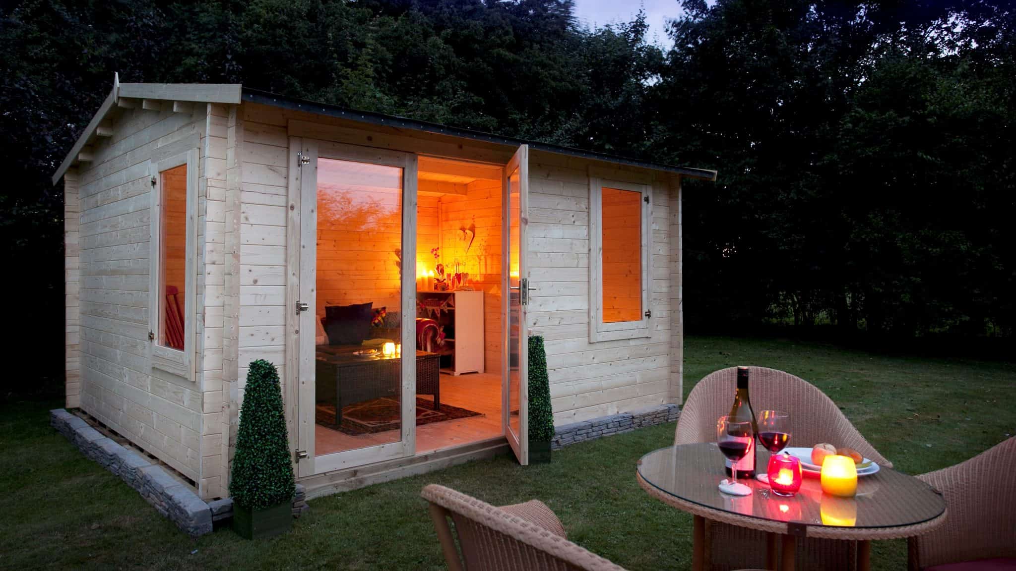 wooden shed summerhouse at the end of a garden in front of a small table with drinks and lights on it with its door open and lit up inside