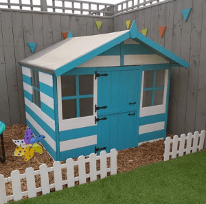 children's blue and white wooden playhouse in a boundary corner next to a fence with small white picket fence border