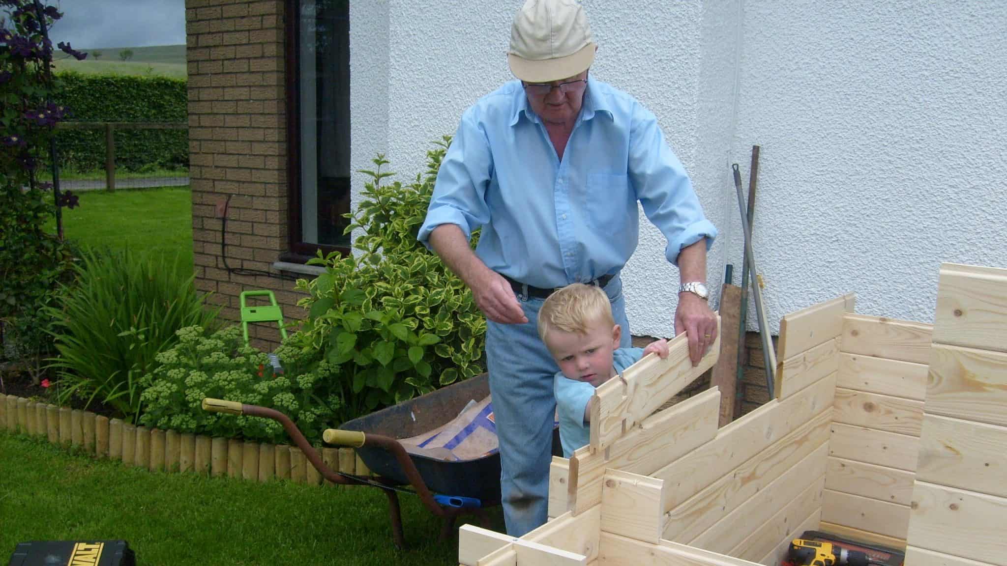 older man in a blue shirt and cap helping a young blonde child build a timber shed wall in the garden